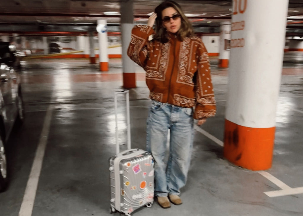 Person in a patterned jacket and jeans standing with a suitcase in a parking garage.